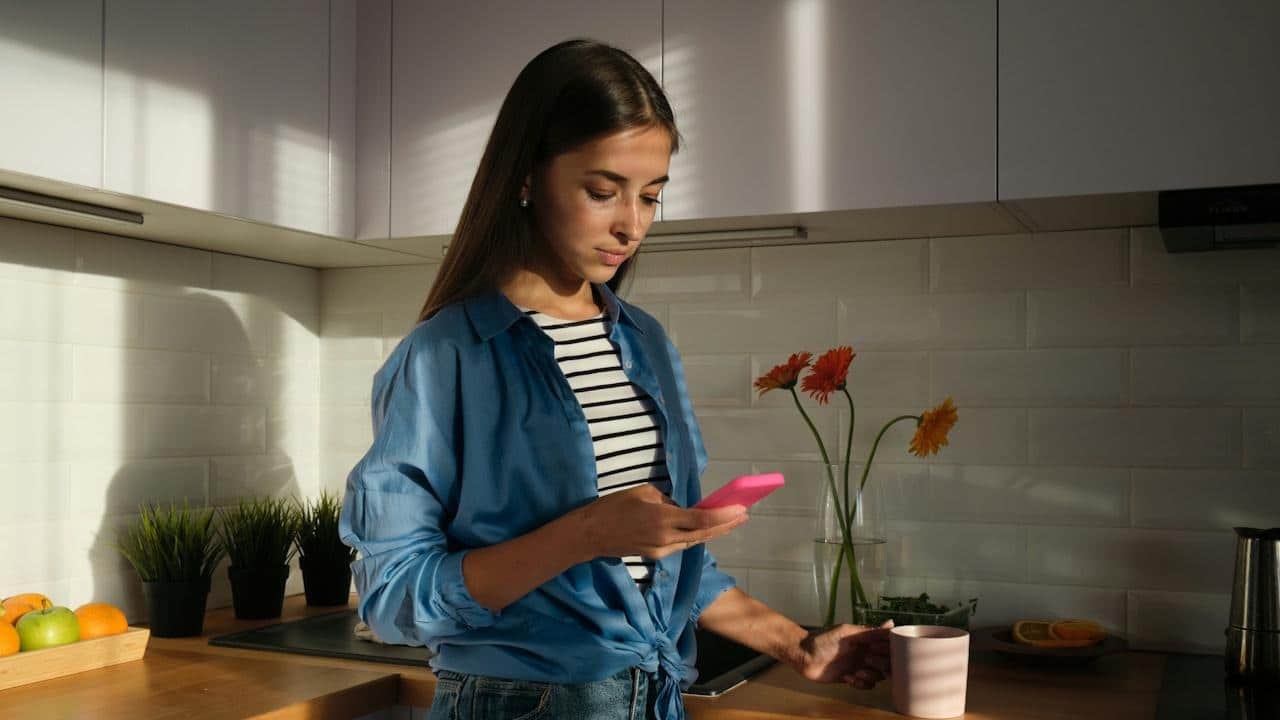 A young woman standing in a kitchen looking at her smartphone while holding a coffee mug.