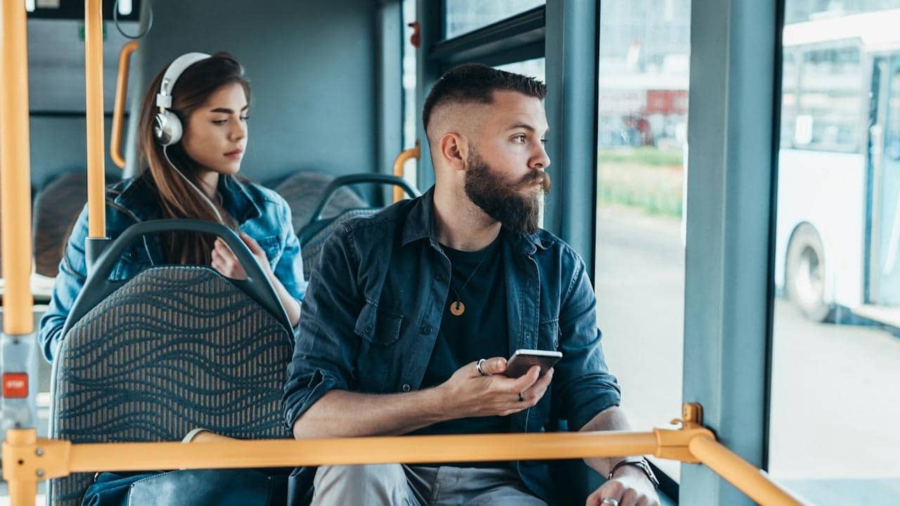 A bearded man wearing a dark shirt and rings looks out the bus window, while a woman in a denim jacket and headphones sits behind him.