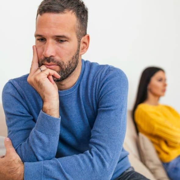 A man in a blue sweater sits looking down while a woman sits blurred behind.