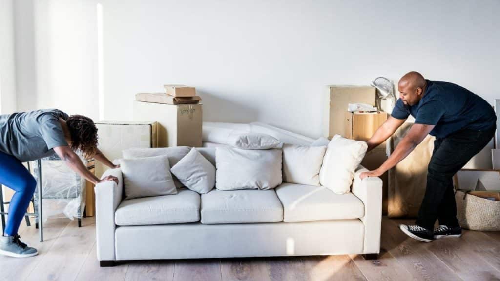 Two people lean over to lift a white sofa in a room filled with boxes.