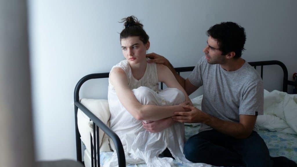 A man touches the shoulder of a woman sitting on a bed looking away.