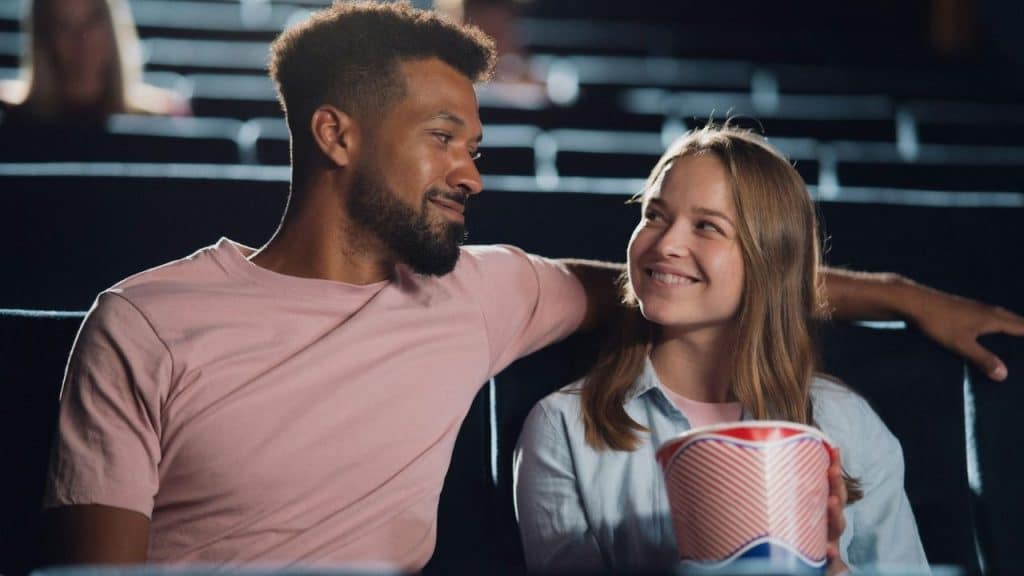 A man and woman sit together in a dark theater, smiling and holding popcorn.