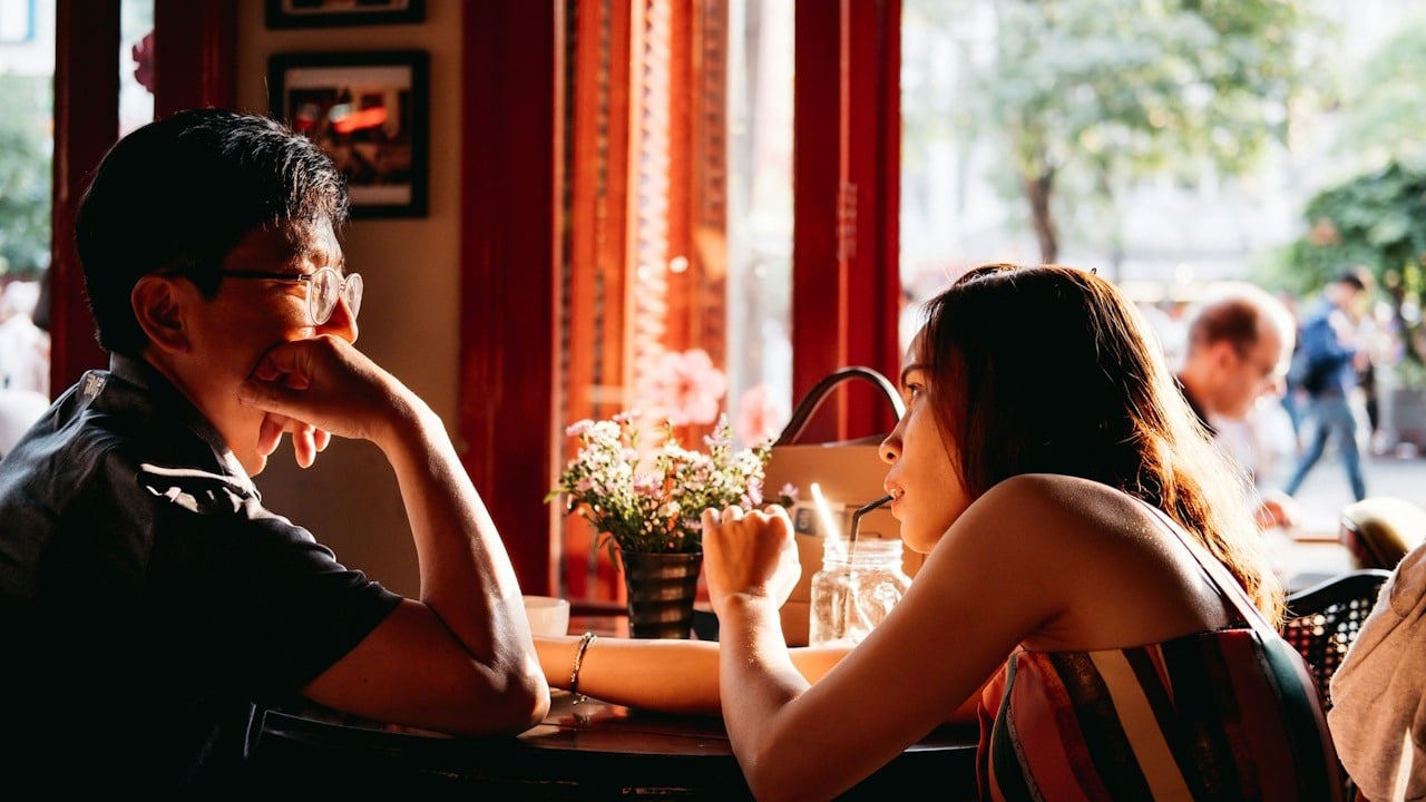 Two people sit across from each other at a small café table by a window, engaged in conversation.