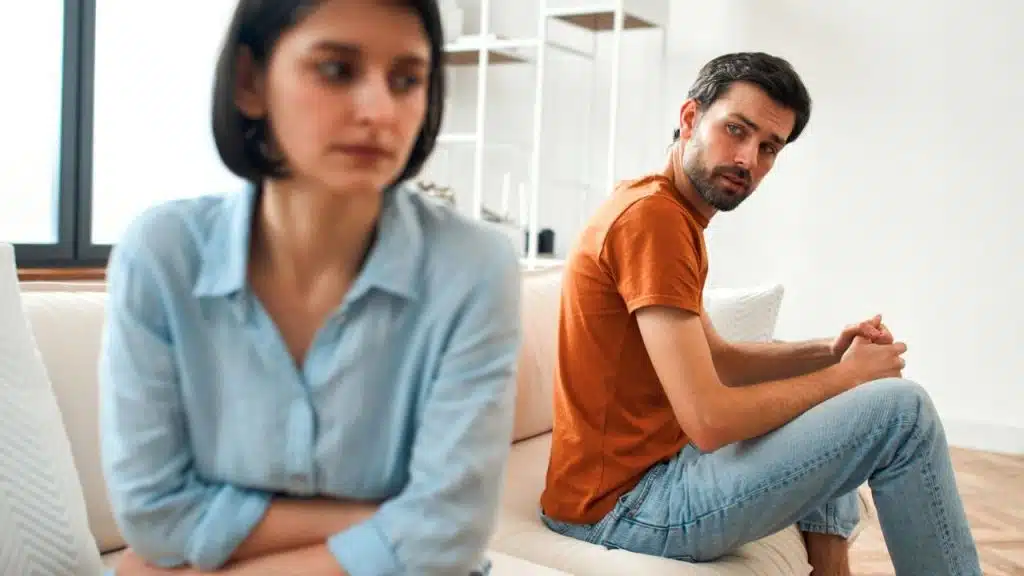 A man in an orange shirt looks toward a woman sitting with her arms crossed.