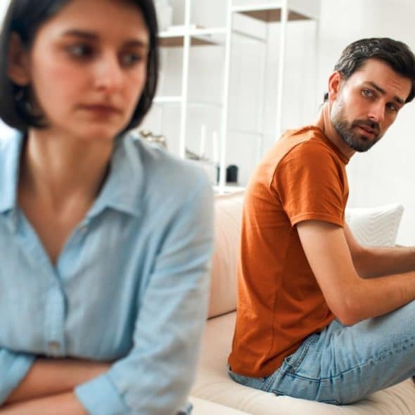 A man in an orange shirt looks toward a woman sitting with her arms crossed.