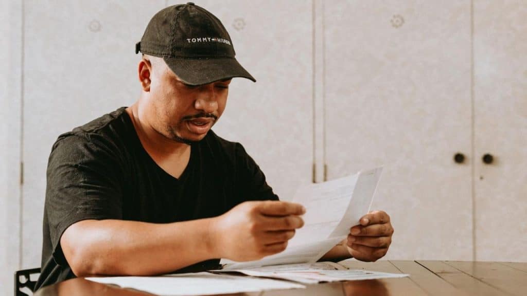 A man wearing a black baseball cap sits at a table while looking at papers.