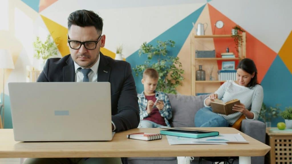 A man works on a laptop while a woman and child sit in the background.