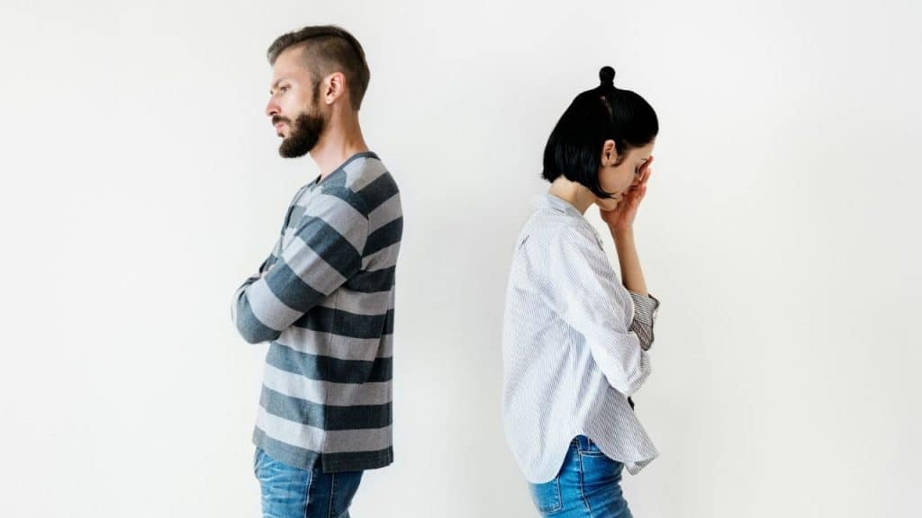 A man and woman stand back-to-back against a white wall with frustrated facial expressions.