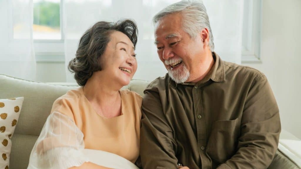 An older man and woman sit on a sofa and smile at each other.