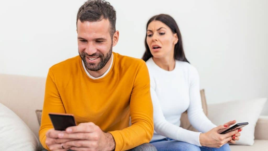 A woman looks over a man's shoulder while they both sit and use mobile phones.