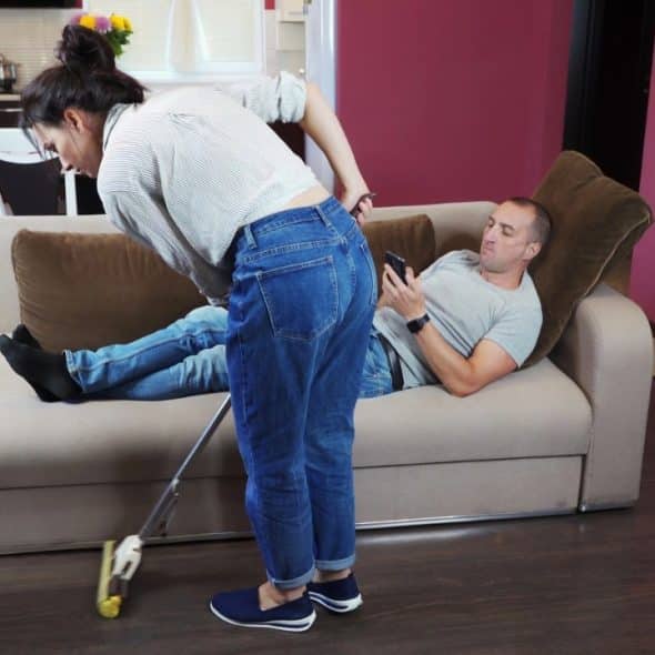 A woman in jeans mops the floor while a man reclines on a nearby sofa.