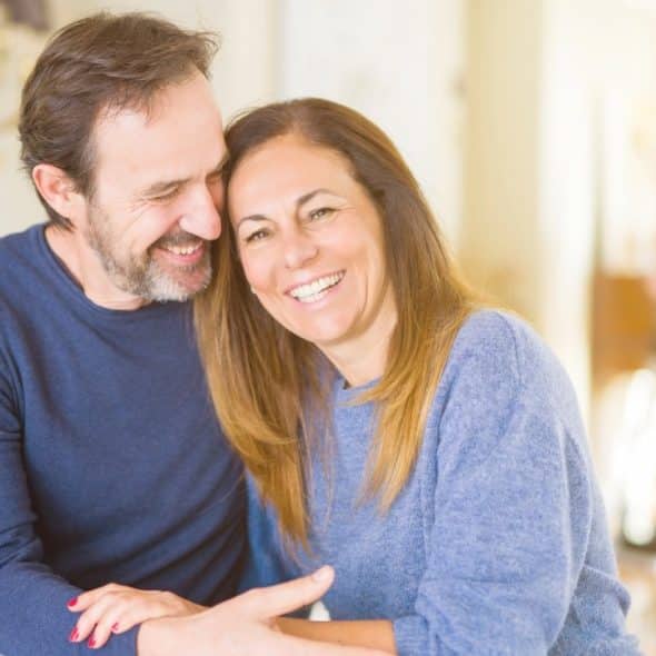 A smiling man and woman sitting close together, leaning their heads toward one another indoors.