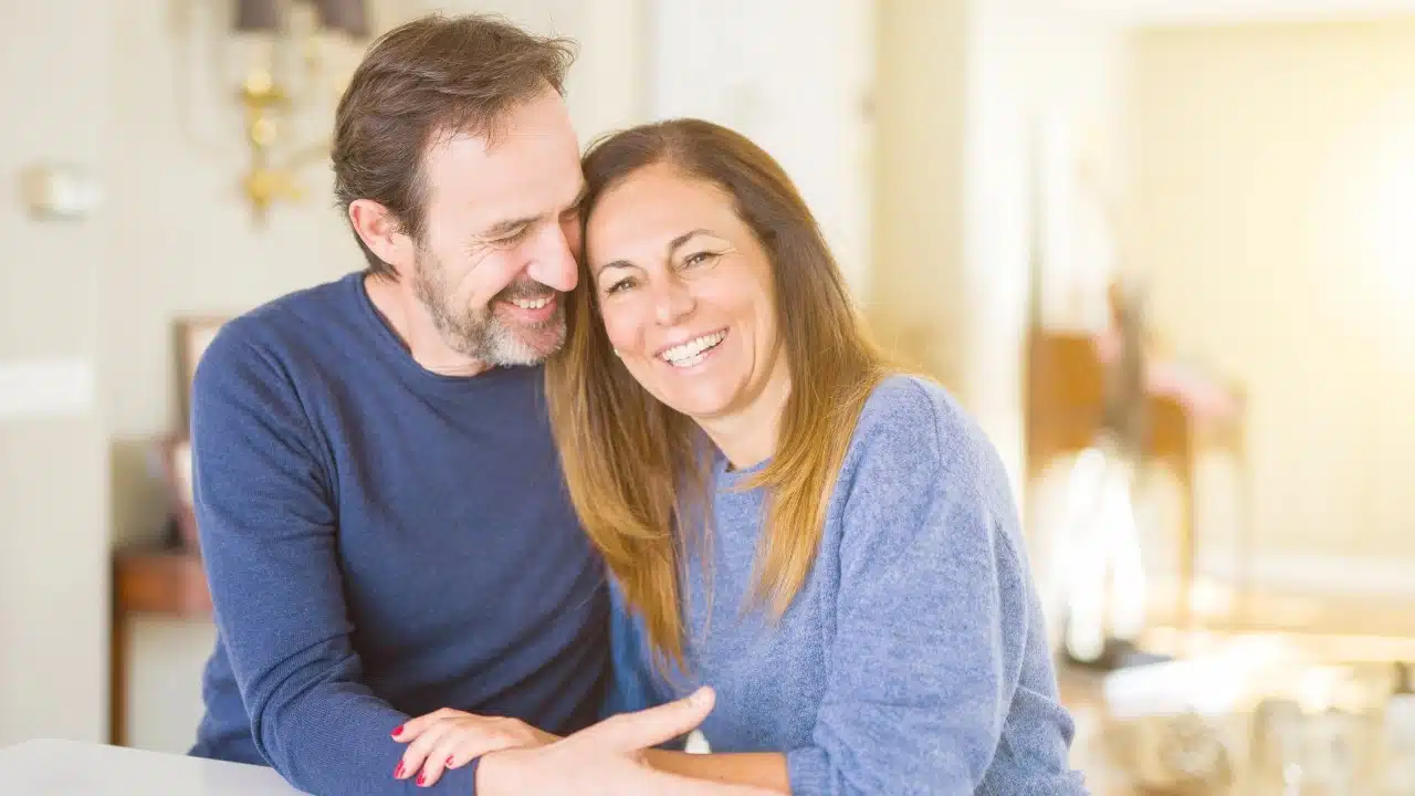 A smiling man and woman sitting close together, leaning their heads toward one another indoors.