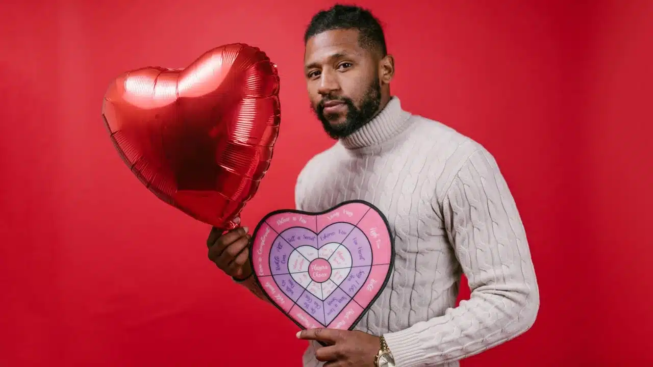 A man in a beige sweater holding a red heart balloon and a heart-shaped board.