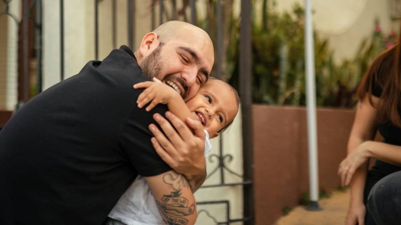 A smiling man with tattoos on his arm giving a small child a tight hug.
