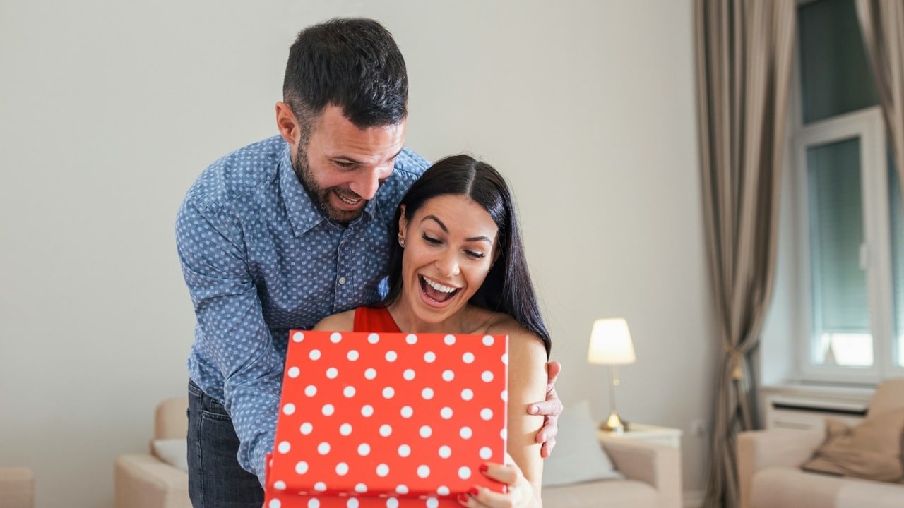 A man and woman smiling while opening a red gift box with white polka dots.