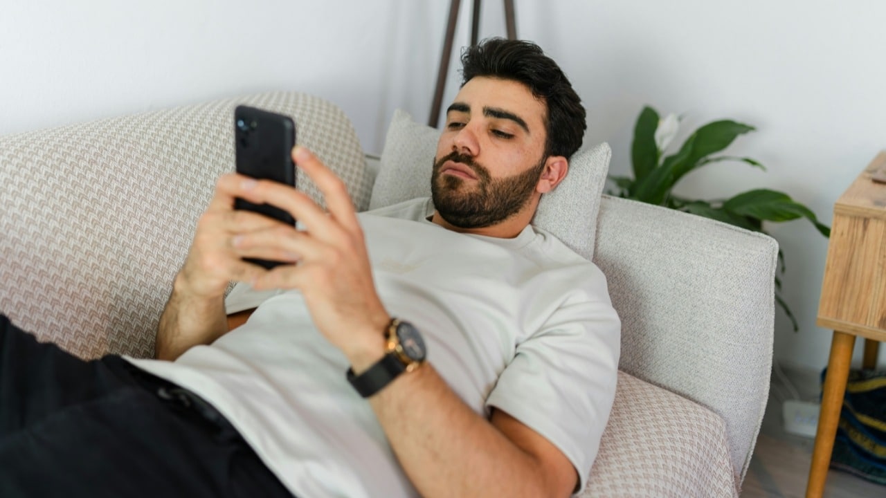 A bearded man reclining on a patterned couch while holding a black smartphone with both hands.