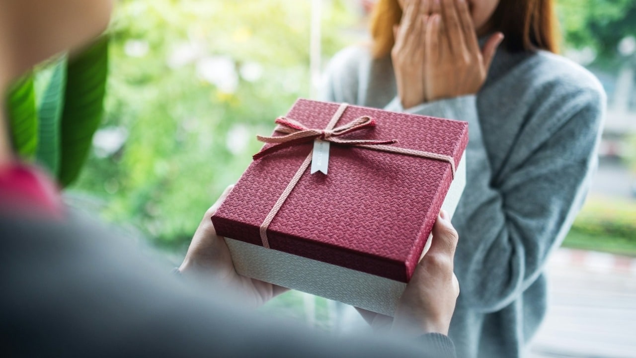 Close-up of hands presenting a red gift box to a person covering their mouth.