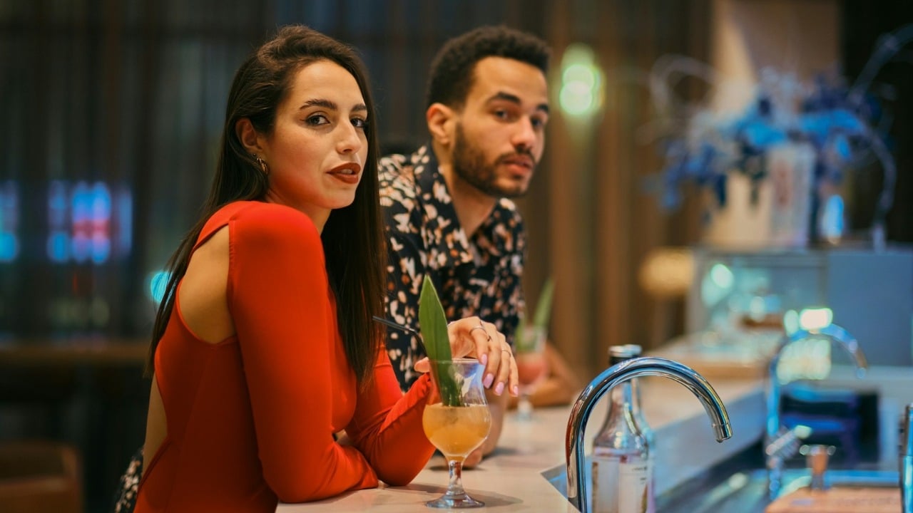 A man and woman sitting at a bar with drinks, looking toward the camera.