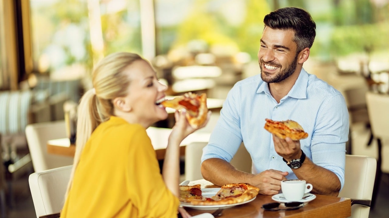 A man and woman sitting at an outdoor table, smiling and laughing while eating large pizza slices.