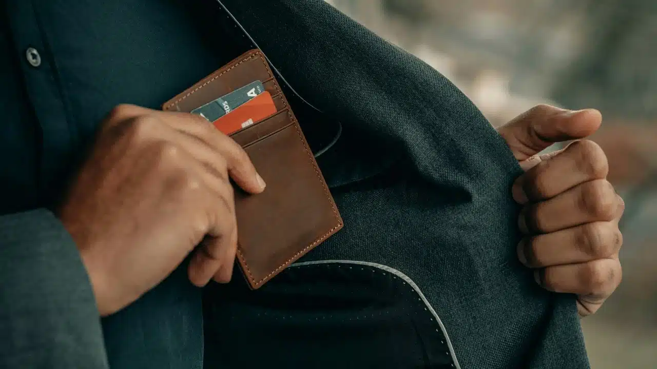 Close-up of a person in a gray suit jacket pulling a brown leather wallet from an inner pocket.