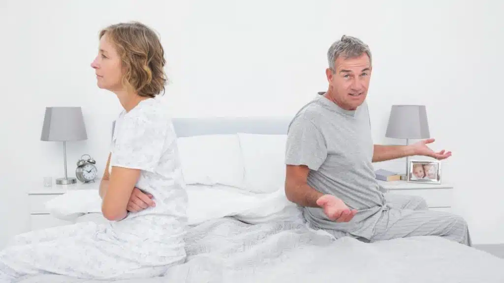 A man and woman sit back-to-back on a bed; the man gestures with open palms.