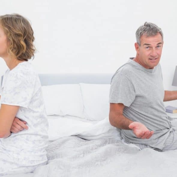 A man and woman sit back-to-back on a bed; the man gestures with open palms.
