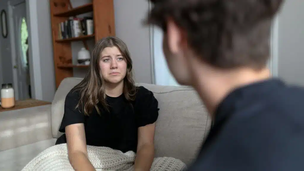 A woman with long brown hair sits on a sofa, looking directly at a person in the foreground.