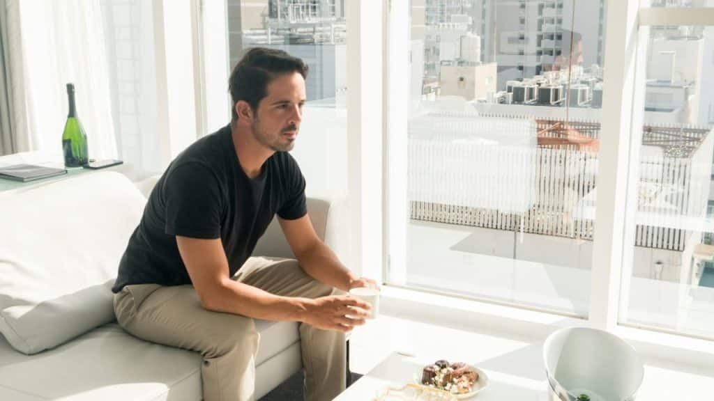 A man in a black shirt sits on a white sofa holding a white mug.