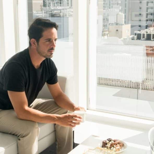 A man in a black shirt sits on a white sofa holding a white mug.