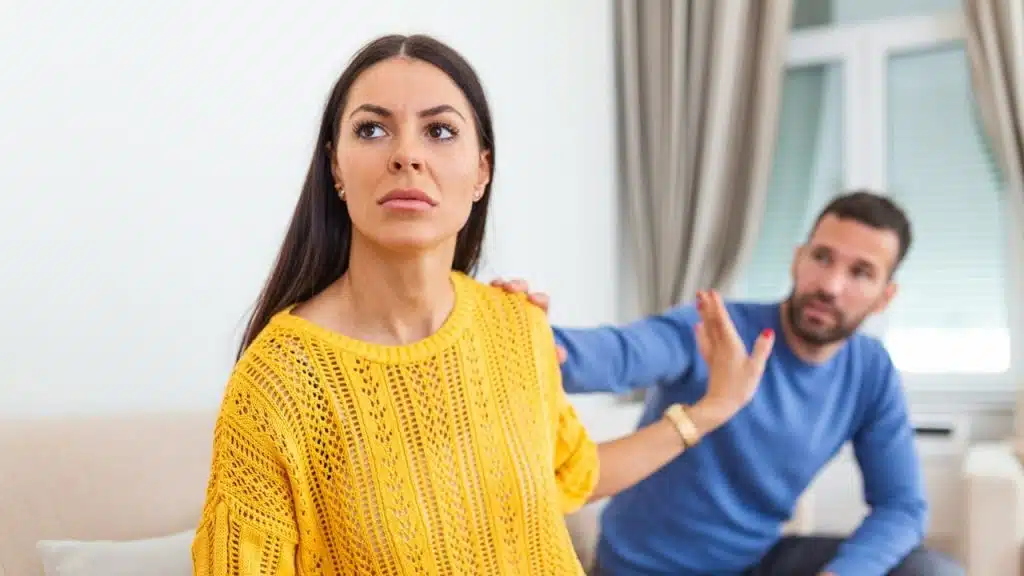 A woman in a yellow sweater looks away while a man reaches toward her shoulder behind her.