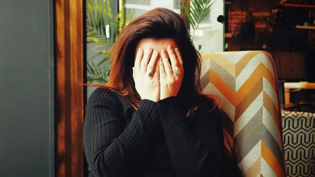 A woman with long reddish-brown hair sits in a chair, covering her face with both hands.