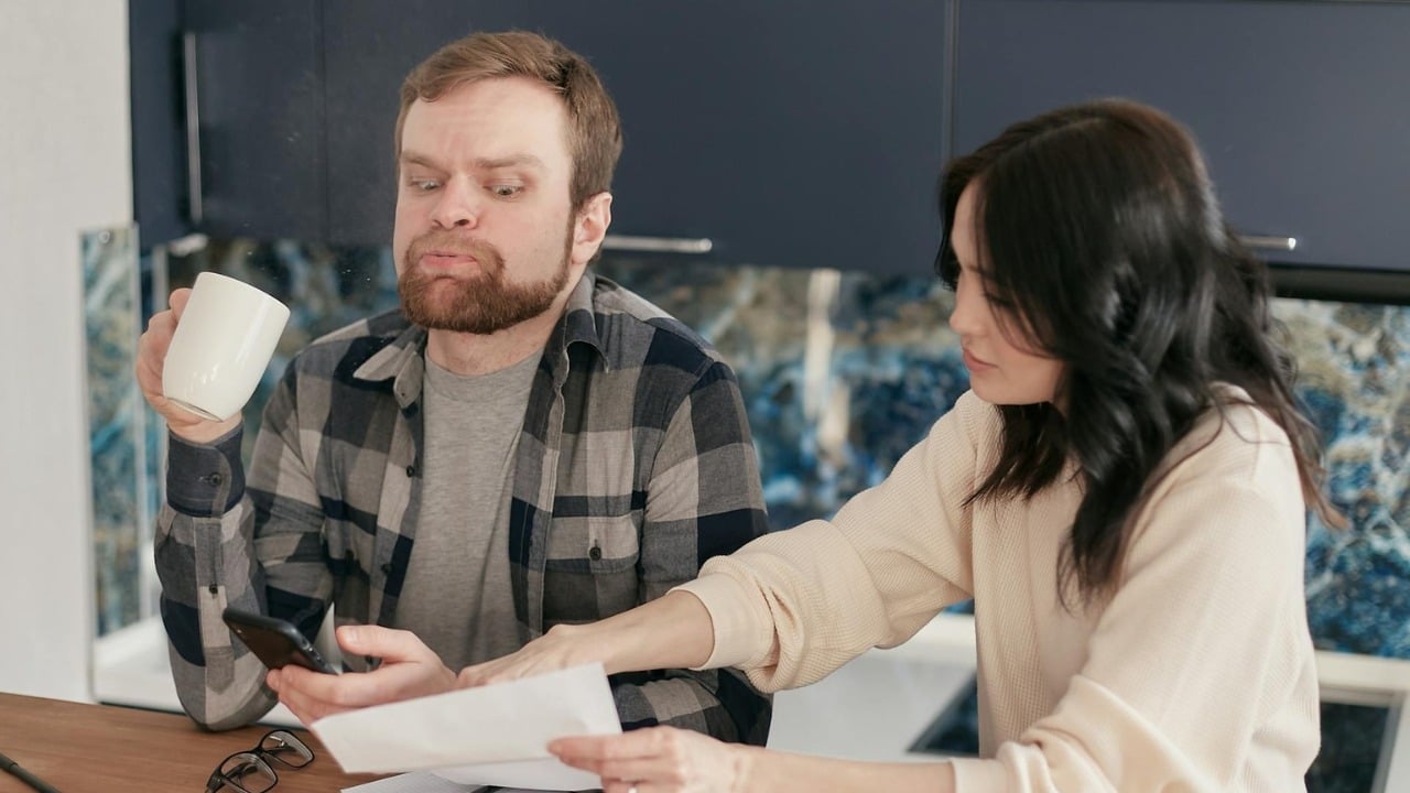 A man and woman sitting at a table looking at a smartphone and paper documents.