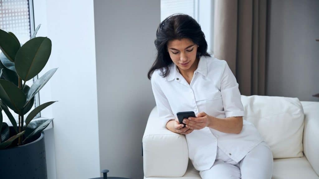 A woman in a white outfit sits on a white couch and looks at a smartphone.