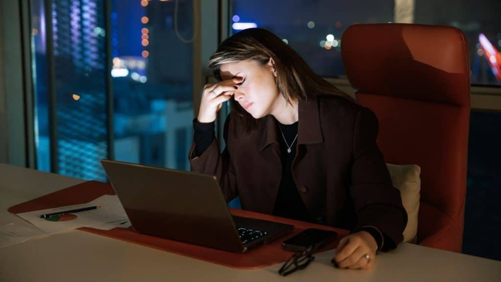 A woman at a desk touches her forehead while looking at a laptop at night.
