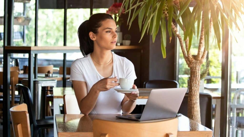 A woman in a white t-shirt sits at a table with a laptop and coffee.