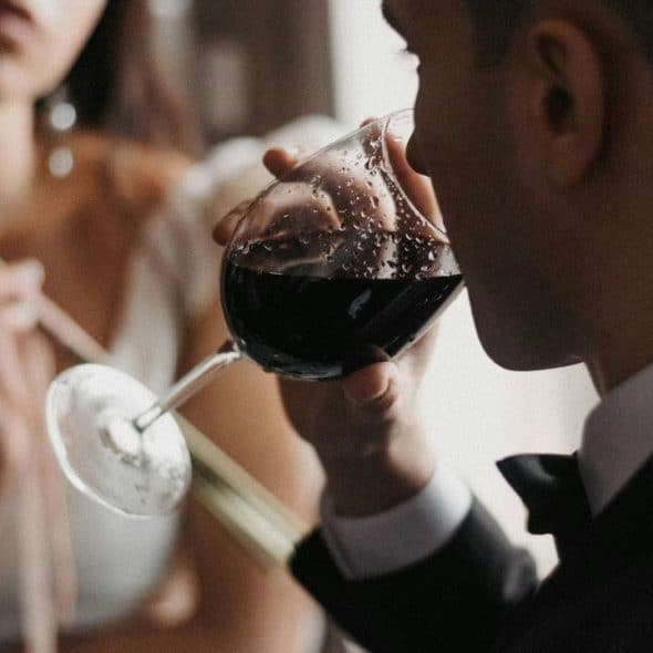 A man in a suit sipping red wine while a woman holds a red flower in the background.