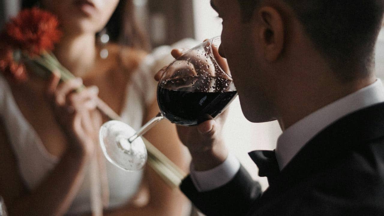 A man in a suit sipping red wine while a woman holds a red flower in the background.