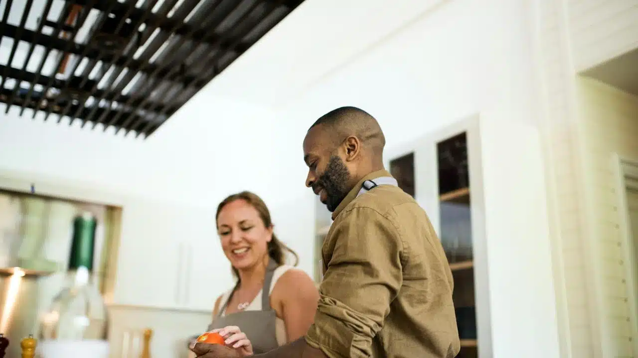 A couple smiling while cooking together in a bright kitchen.