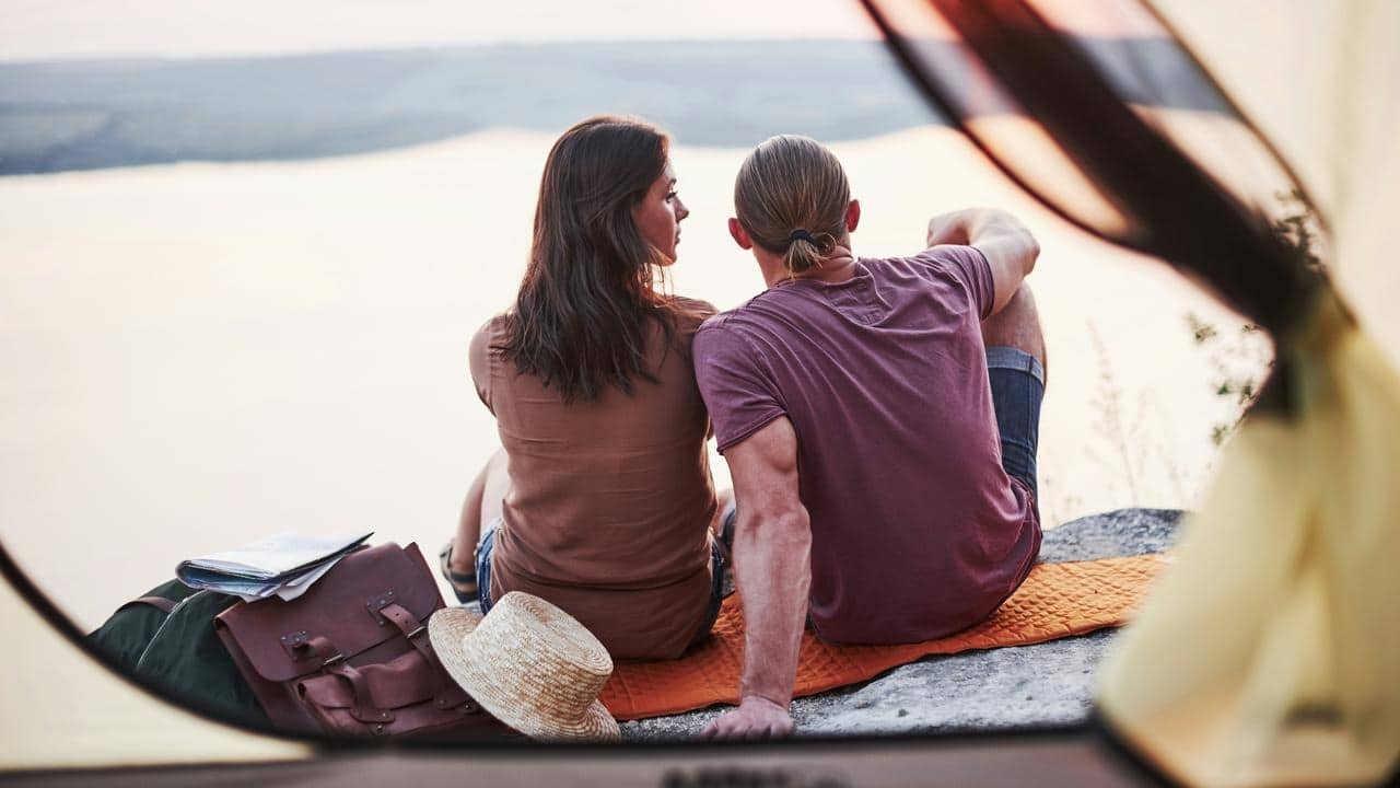 A couple sitting on a blanket by the water, viewed from inside a tent.