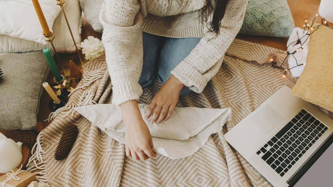A person folding fabric on a cozy blanket beside a laptop and candles.
