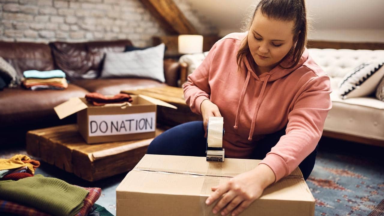 A woman sealing a cardboard box with tape beside a donation box of clothes.