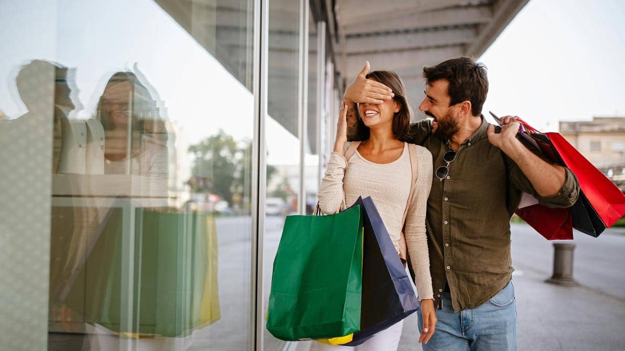 A man playfully covering a woman’s eyes while they carry shopping bags outside a store.