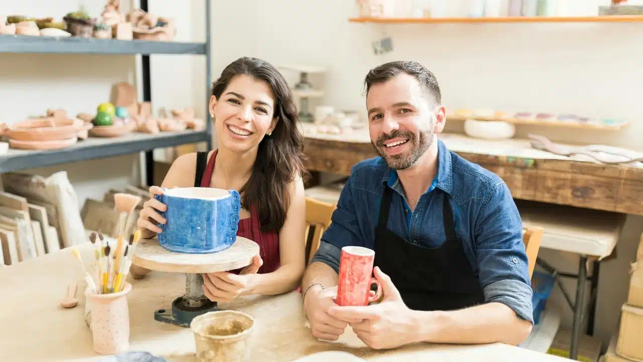 A couple smiling in a pottery studio while holding handmade ceramic pieces.