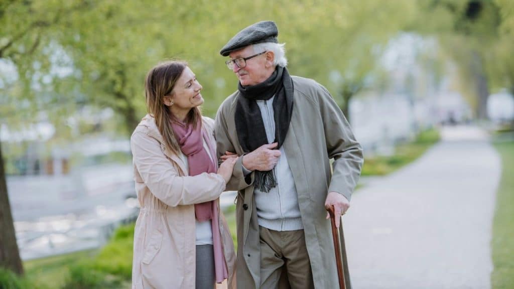 A woman walks alongside an older man with a cane on a tree-lined path.