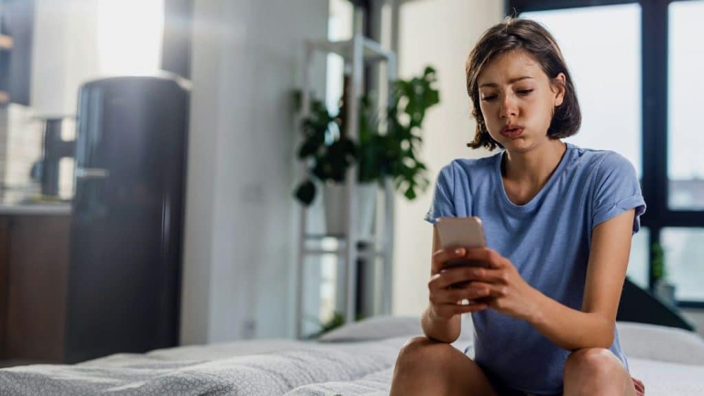 A young woman sits on a bed with a sad expression while looking at a smartphone.