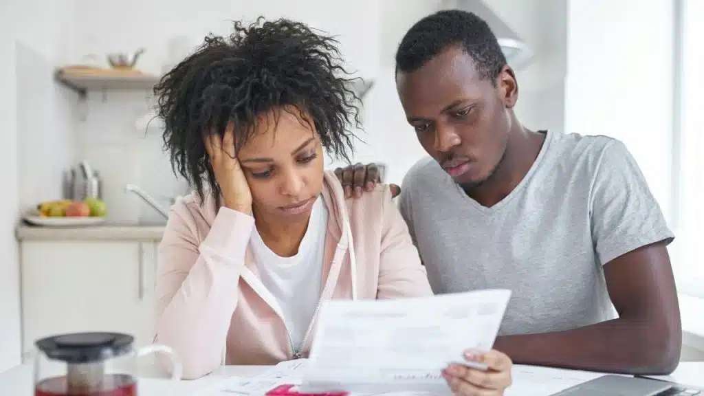 A man and woman sit at a table looking at a document with concerned expressions.