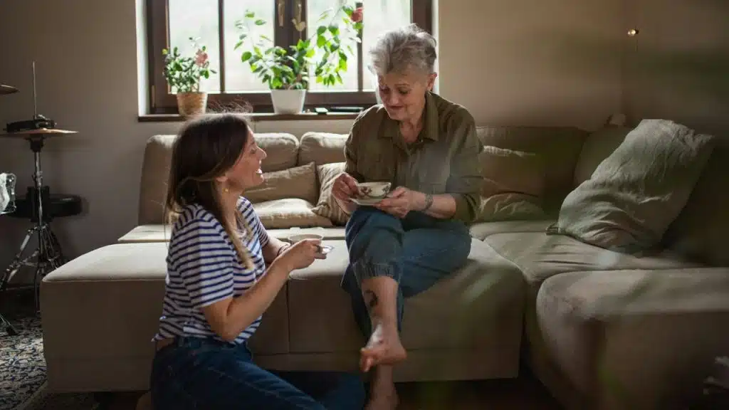 Two women sit together on and near a couch, smiling and holding tea cups.