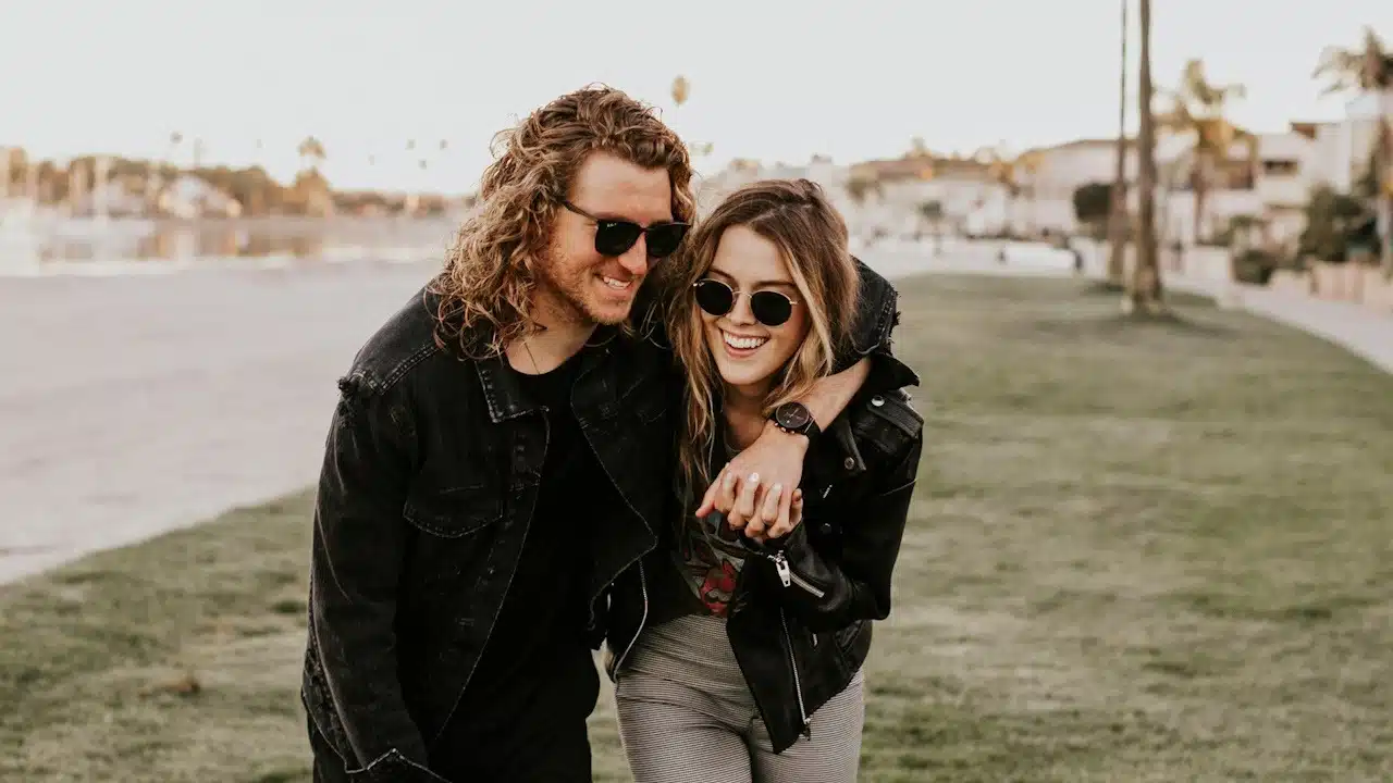 Man dressing black hugging a woman in black leather jacket and gray pants on green grass field during daytime.