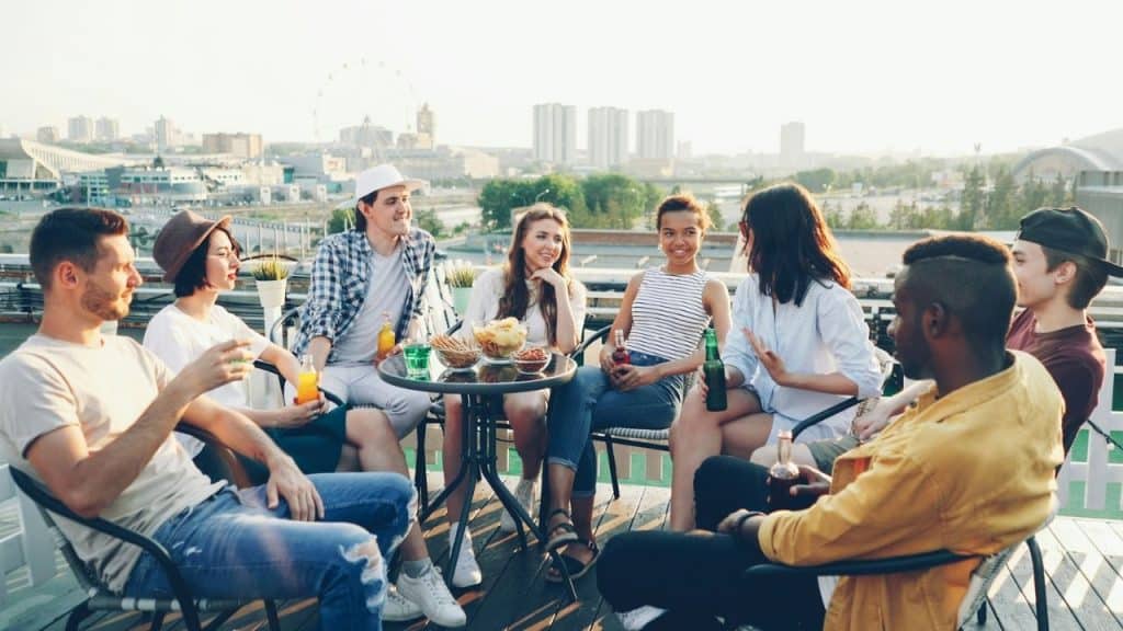 A group of young adults sits around a table on a rooftop patio with drinks.
